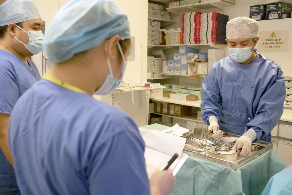 A picture of three staff members in a theatre. One of the theatre staff is preparing medical equipment ready for surgery