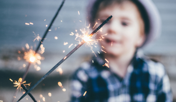 An image of lit sparklers. In the background is a child stood outside, he is looking at the sparklers.